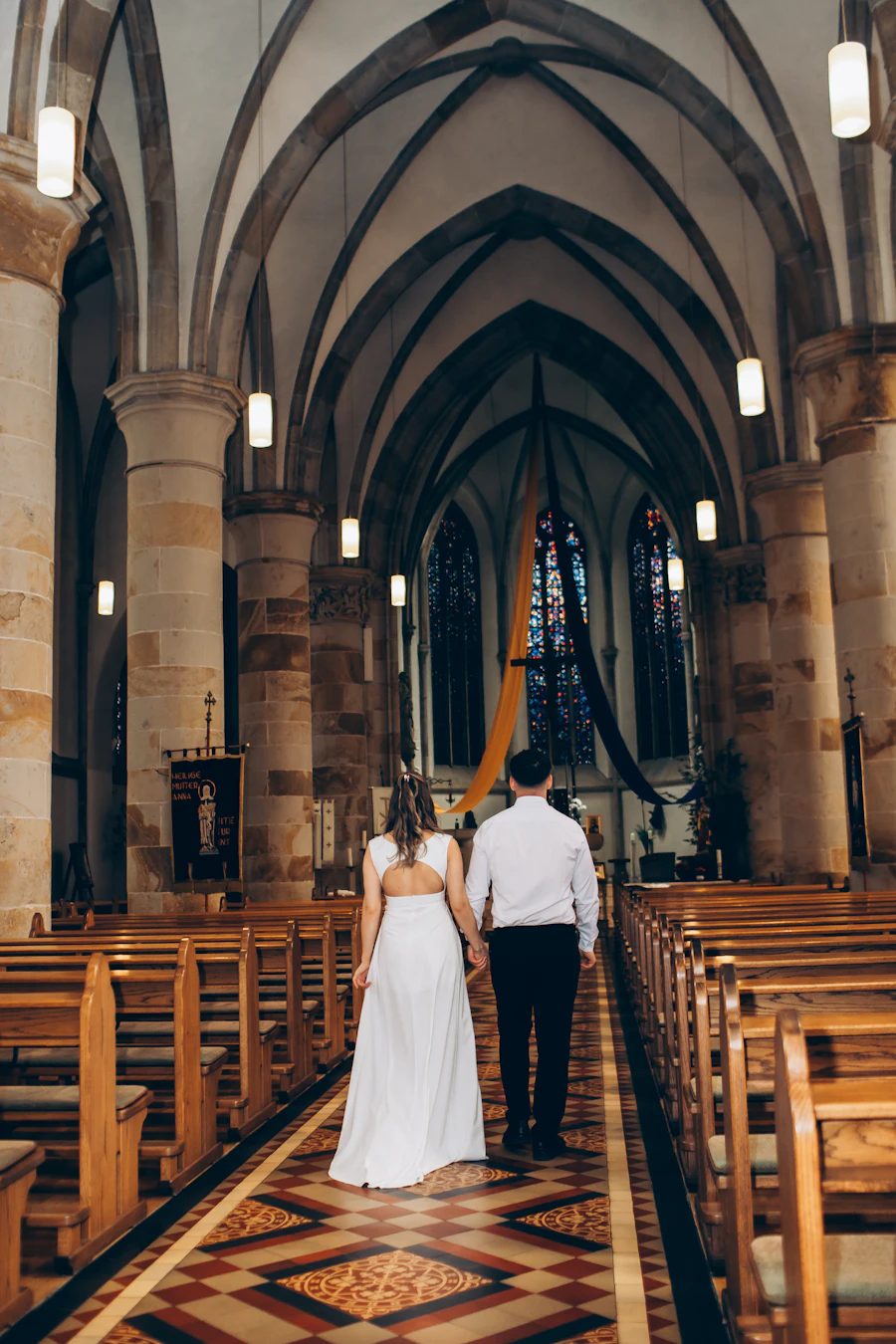A bride and groom walking hand in hand down a church aisle, seen from behind