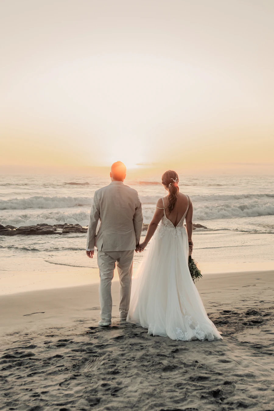 A bride and groom walking hand in hand on the beach toward the sunset, seen from behind
