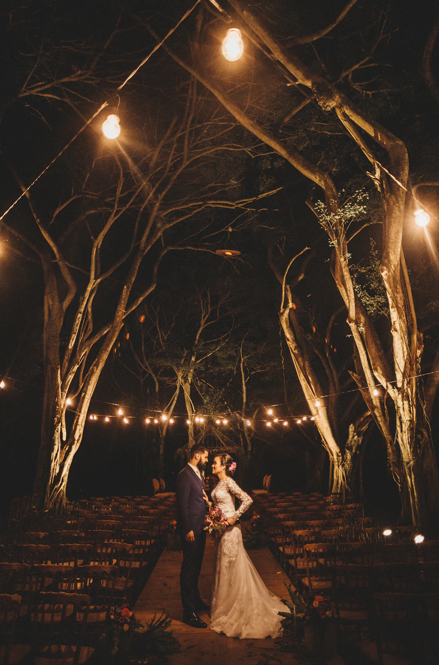 A bride and groom standing under trees hung with warm string lights at night