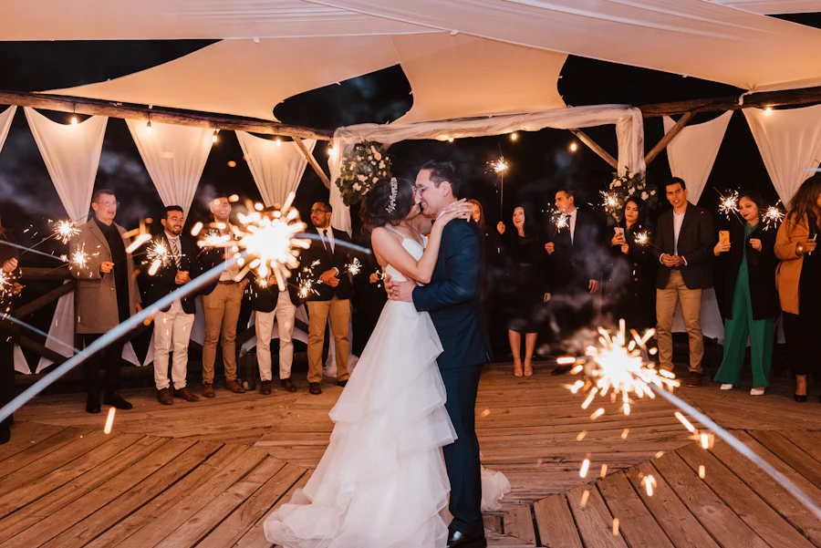 A bride and groom kissing while guests wave sparklers around them