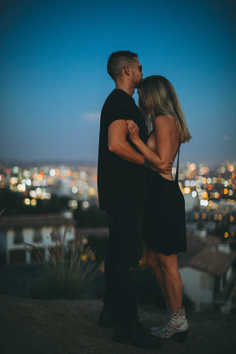 A man hugging a woman on a hilltop at night, with city lights below